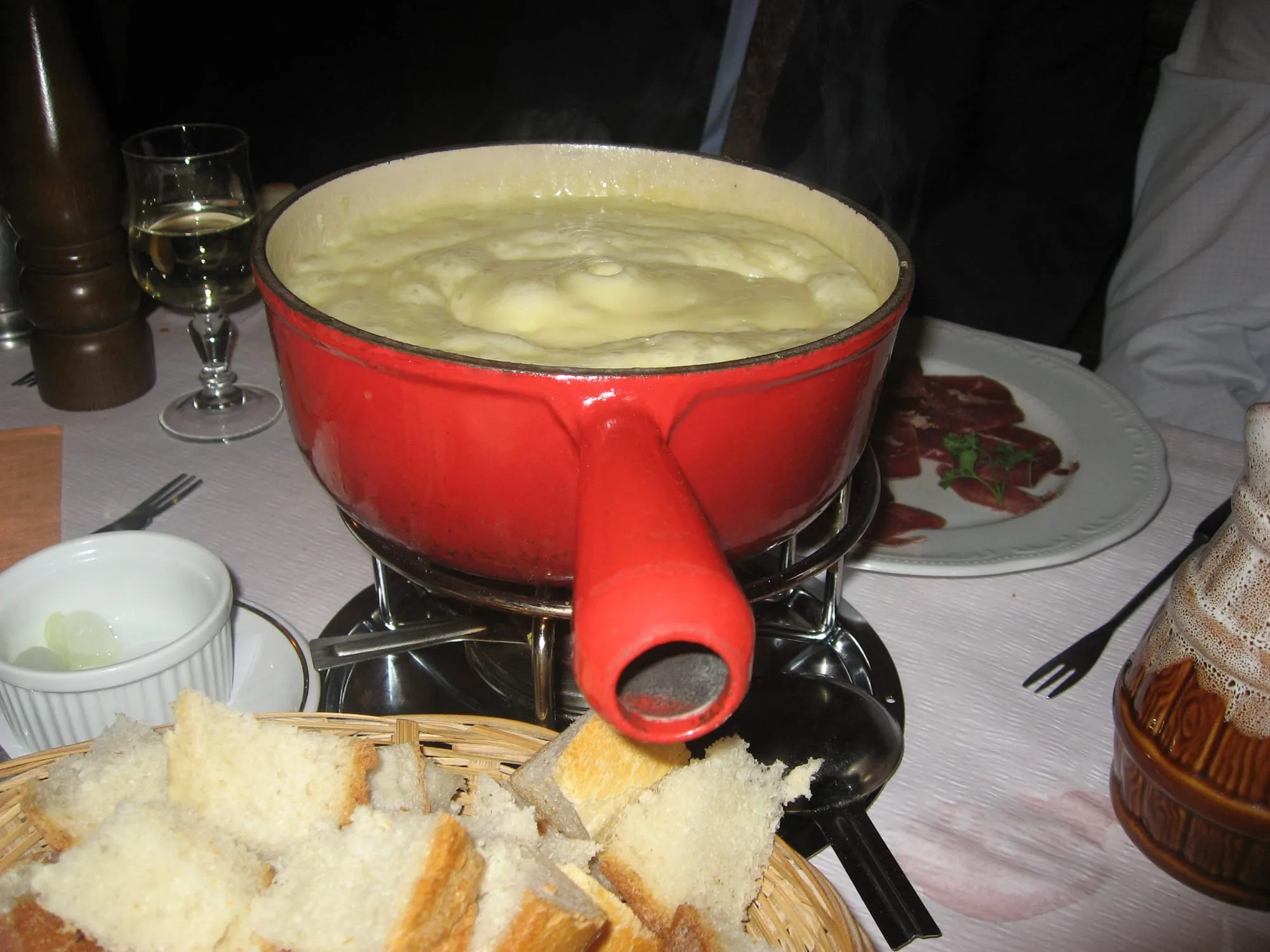 Una cassola vermella de fondue plena de formatge fos que fumeja damunt del fogonet, en una taula de restaurant parada amb una cistella de pa a trossos i un plat de carn seca.